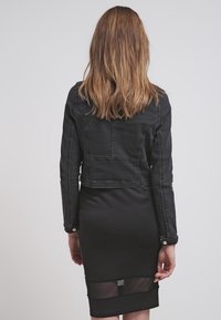 Veste en denim noir à coupe ajustée, avec des coutures visibles et des éléments en métal, associée à une robe crayon noire avec un détail d'ourlet en tissu transparent.
