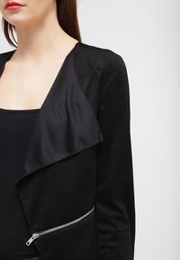 Veste noire avec une texture douce rappelant le daim. Elle présente un col drapé et un détail de zip argenté sur le côté, conçue pour une silhouette ajustée.