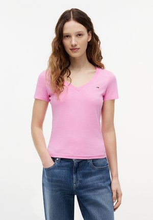 Junge Frau mit langen, welligen Haaren, trägt ein rosafarbenes V-Ausschnitt-T-Shirt und blaue Jeans, steht mit einer Hand in der Tasche vor weißem Hintergrund.