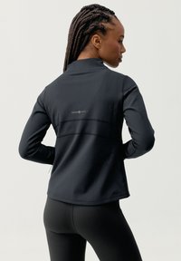 Czarna sportowa kurtka z wysokim kołnierzem, dopasowanym krojem i fakturowanymi panelami z tyłu. Posiada logo na górnej części pleców.