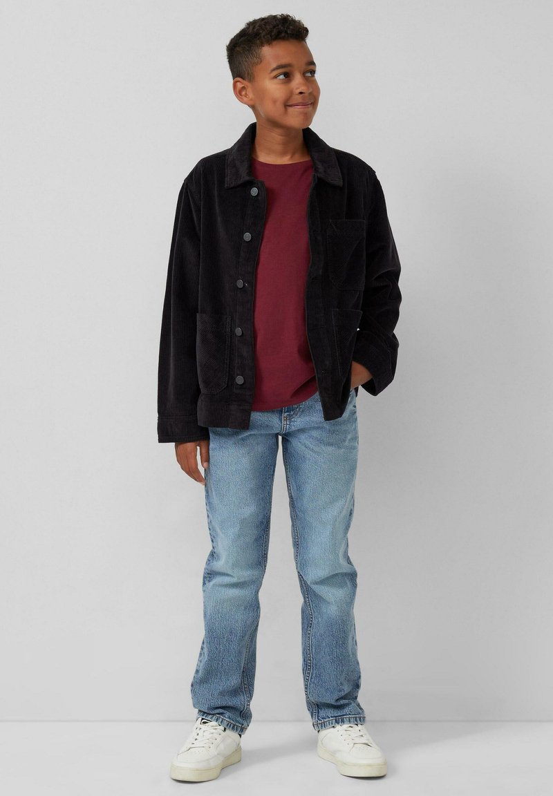 Dunkel schwarze Cordjacke mit Knöpfen, getragen über einem bordeauxroten T-Shirt, kombiniert mit hellblauen Jeans und weißen Sneakers.