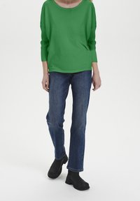 Zielony sweter z prążkowanego dzianiny o szerokim dekolcie, zestawiony z prostymi niebieskimi dżinsami i czarnymi botkami za kostkę. Prosty i swobodny strój.