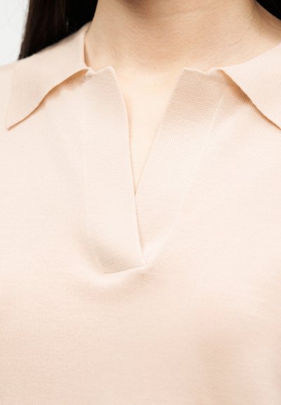 Femme portant une chemise beige clair à col en V avec une texture de tissu douce et lisse visible autour du cou et de la partie supérieure de la poitrine.
