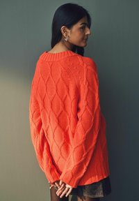 Übergroßer oranger Strickpullover mit einem Rautenmuster und geripptem Halsausschnitt, ausgestattet mit langen Ärmeln und einer lockeren Passform.