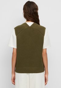 Oliwkowa dzianinowa kamizelka z dekoltem w serek i żebrowanym wykończeniem na ramionach oraz na dole. Noszona na jasnym t-shircie.