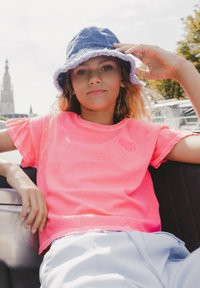 Roze T-shirt met korte mouwen en een rafelige zoom, voorzien van een logo. Draagt een blauw denim bucket hat met een rafelige rand en decoratieve stiksels.