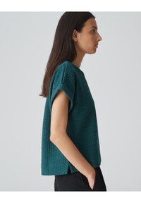 Teal textuur top met korte mouwen, met een relaxte pasvorm, zijslits en een subtiel geometrisch patroon. Draagt met zwarte broek.