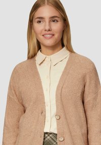 Cardigan marrone chiaro con un tessuto morbido e texture, apertura frontale e tre grandi bottoni. Indossato sopra una camicia bianca a bottoni.