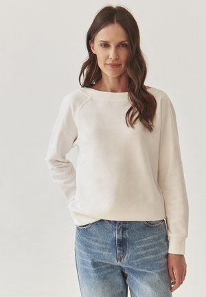 White sweatshirt with a boat neckline and ribbed cuffs, paired with light blue faded jeans. The fabric appears soft and textured.