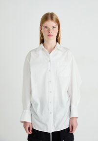 Jongere met steil blond haar draagt een oversized wit overhemd met knopen en grote oorringen, staand tegen een effen achtergrond.