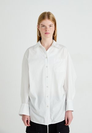 Jongere met steil blond haar draagt een oversized wit overhemd met knopen en grote oorringen, staand tegen een effen achtergrond.
