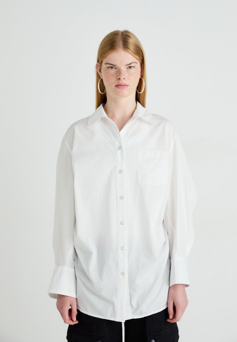 Jongere met steil blond haar draagt een oversized wit overhemd met knopen en grote oorringen, staand tegen een effen achtergrond.