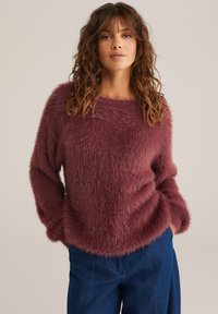 Fluffy, kastanjebruine trui met lange mouwen en een wijde halslijn; gecombineerd met losse blauwe jeans, die een zachte textuur en ontspannen vorm laten zien.