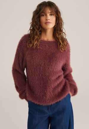 Fluffy, kastanjebruine trui met lange mouwen en een wijde halslijn; gecombineerd met losse blauwe jeans, die een zachte textuur en ontspannen vorm laten zien.