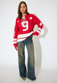 Sudadera roja con el número 9 en blanco y acentos de estrellas, combinada con jeans de mezclilla azules de pierna ancha con detalles de patchwork.