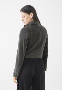 Chaqueta recortada de cuero verde oscuro con cuello, puños con cremallera y textura sutil. Llevada sobre una prenda negra con plisados.