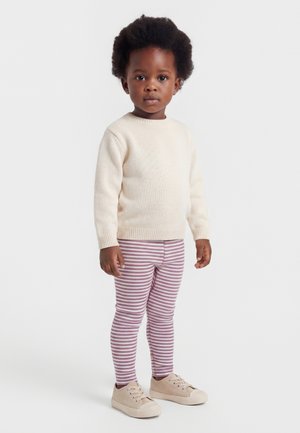 Väikelaps lokkis juustega, kandes kremi värvi kampsunit, roosa-valgete triibuliste retuusidega ja beeže vabaajajalatsitega, seisab ühevärvilise tausta ees.