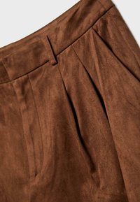 Pantaloni in suede marrone in grande dettaglio che mostrano la parte anteriore plissettata, la vita con passante per la cintura e il tessuto morbido e testurizzato.
