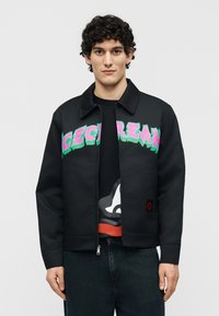 Νέος άνδρας με κοντά σγουρά μαλλιά, που φοράει μαύρο τζάκετ με φερμουάρ, με ροζ και πράσινο κείμενο στο στήθος, και σκούρο τζιν.