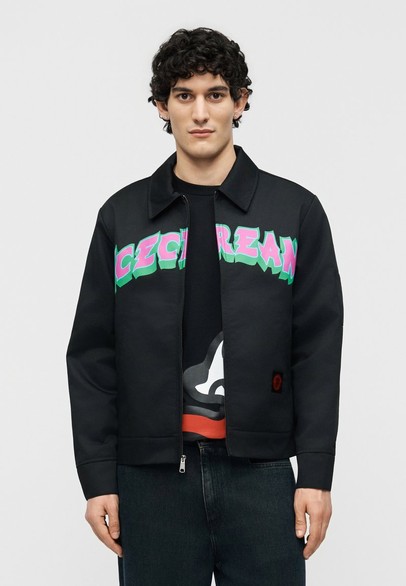 Νέος άνδρας με κοντά σγουρά μαλλιά, που φοράει μαύρο τζάκετ με φερμουάρ, με ροζ και πράσινο κείμενο στο στήθος, και σκούρο τζιν.