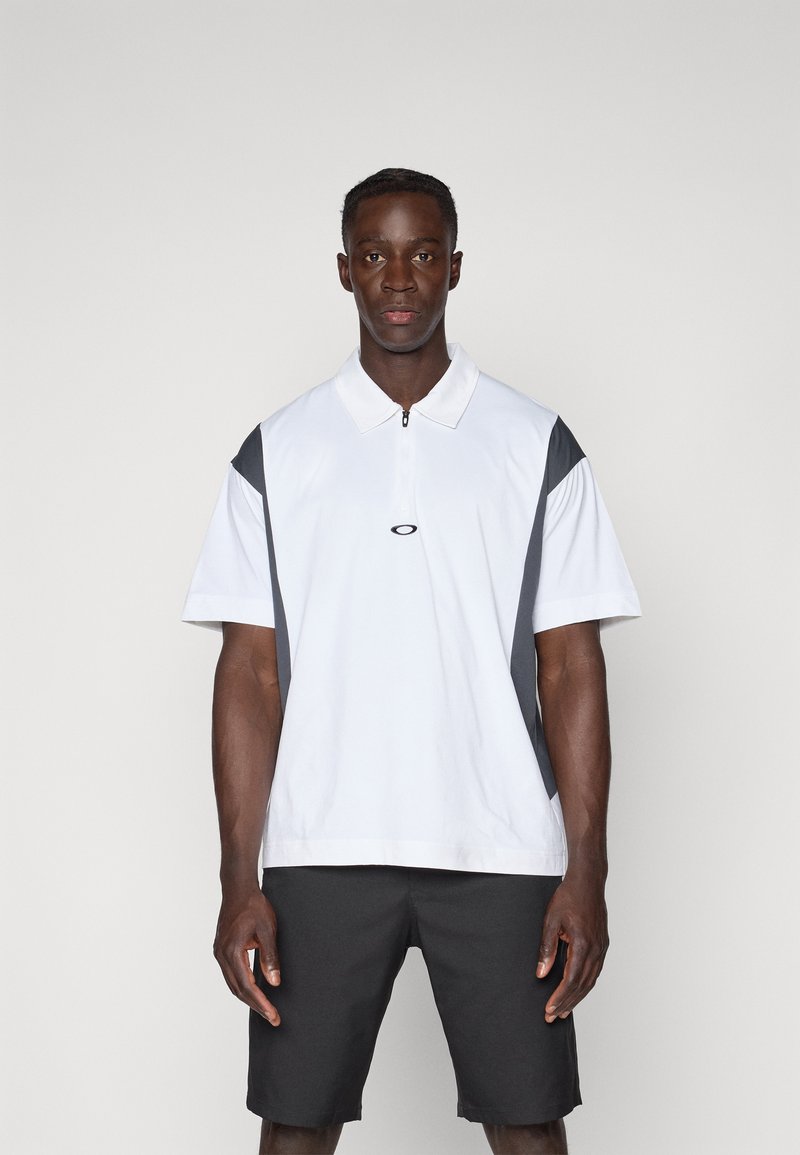 Kortermet polo skjorte i hvit, med grå sidelapper, krage og glidelås. Brukt med mørke shorts. Glatt tekstur.