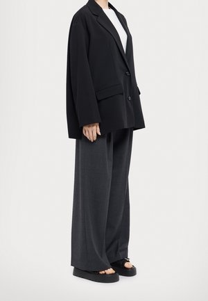 Personne portant un blazer noir surdimensionné, un pantalon large gris et des sandales noires à plateforme ouvertes sur un fond blanc.