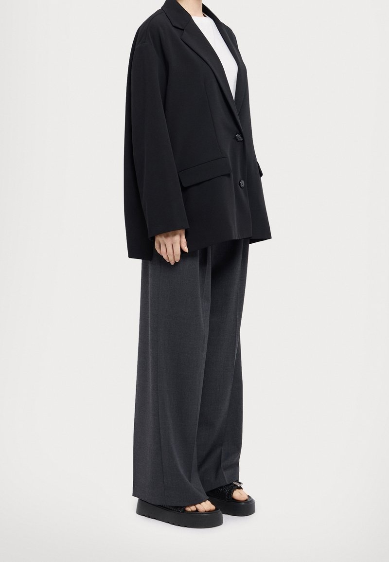 Personne portant un blazer noir surdimensionné, un pantalon large gris et des sandales noires à plateforme ouvertes sur un fond blanc.