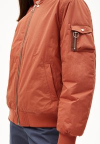 Bomber arancione con zip frontale, polsini a costine e tasca laterale con zip. Texture liscia con una finitura leggermente arricciata.