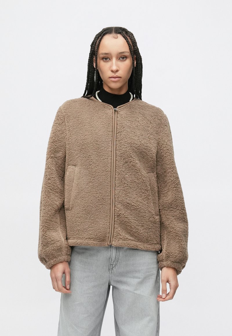 Bruine, pluizige fleecejas met een rits aan de voorkant, geribde kraag en zijzakken. Heeft een ontspannen pasvorm en elastische manchetten.