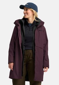 Bordeauxrote wasserdichte Jacke mit Kapuze, Reißverschluss vorne und dunklen Akzenten. Getragen über einem schwarzen Fleecepullover, kombiniert mit braunen Hosen.