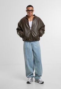 Giubbotto bomber in pelle marrone con zip, jeans in denim azzurro chiaro larghi e sneakers grigie. Caratterizzato da una vestibilità oversize e due tasche frontali.