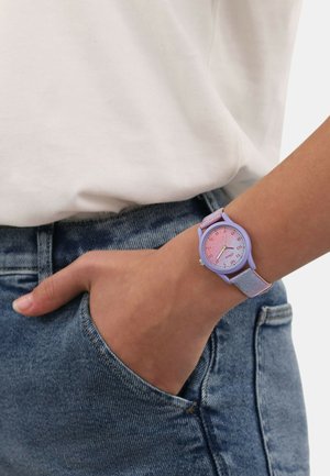 Lila Armbanduhr mit einem glitzernden Armband. Rundes Zifferblatt mit einem pinkfarbenen Verlaufshintergrund und klaren Ziffern. Die Hand ruht in einer Tasche.