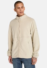 Ljust beige fleecjacka med ståkrage, dragkedja fram, sidofickor och logotypdetalj på vänster ärm. Mjuk, texturerad tyg.