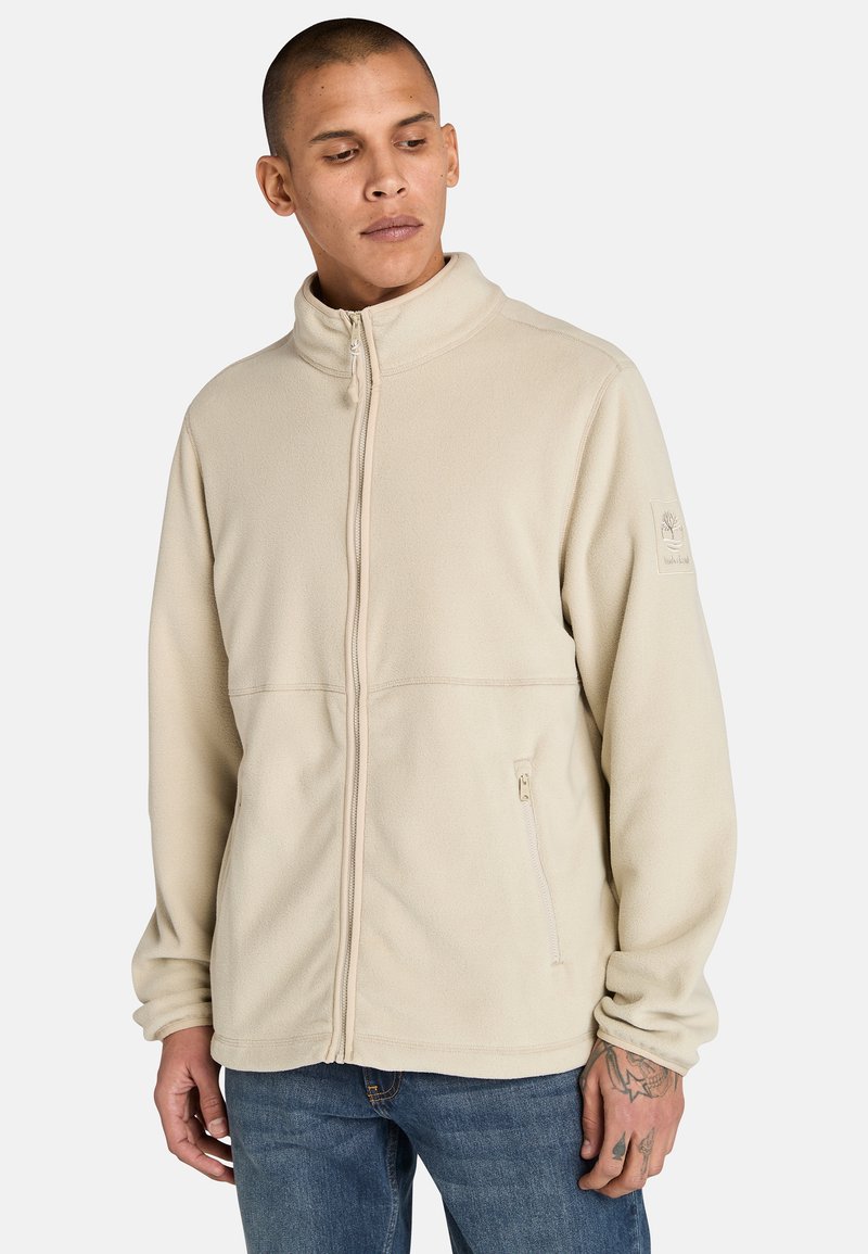 Ljust beige fleecjacka med ståkrage, dragkedja fram, sidofickor och logotypdetalj på vänster ärm. Mjuk, texturerad tyg.
