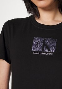 Camiseta de algodón negra con un gráfico rectangular que presenta un fondo gris texturizado y el logotipo "CK" sobre "Calvin Klein Jeans" en texto blanco.
