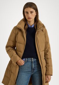 Barna puffermellény magas gallérral, steppelt text�úrával és cipzáros záródással. Egy tengerészkék pulóver és kék farmernadrág fölött viselve.