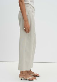 Persona che indossa pantaloni larghi color crema, sandali beige con tacco e un anello d'argento sulla mano sinistra, su uno sfondo bianco semplice.