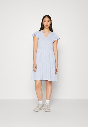 Vestido de algodão azul claro com mangas curtas e babados, decote em V e saia em camadas. Usado com ténis bege e meias brancas até ao tornozelo.