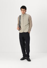 Beige gesteppte Weste mit strukturierten braunen Ärmeln, Reißverschluss vorne und seitlichen Taschen, kombiniert mit schwarzen Hosen und schwarzen Schuhen.