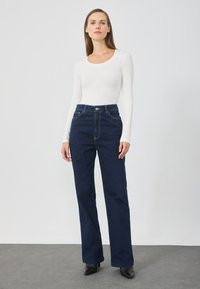 Jeans blu scuro a vita alta con taglio dritto, abbinati a un top bianco a maniche lunghe. Presentano cuciture visibili e una chiusura frontale con bottone.