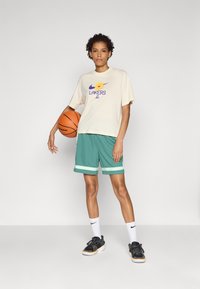 Šviesi grietinėlės spalvos marškinėliai su purpuriniu "Lakers" logotipu ir geltonu gėlių grafiku, derinami su žaliais šortais, kuriuose yra baltos ir žalios juostelės, laikydamas krepšinį.
