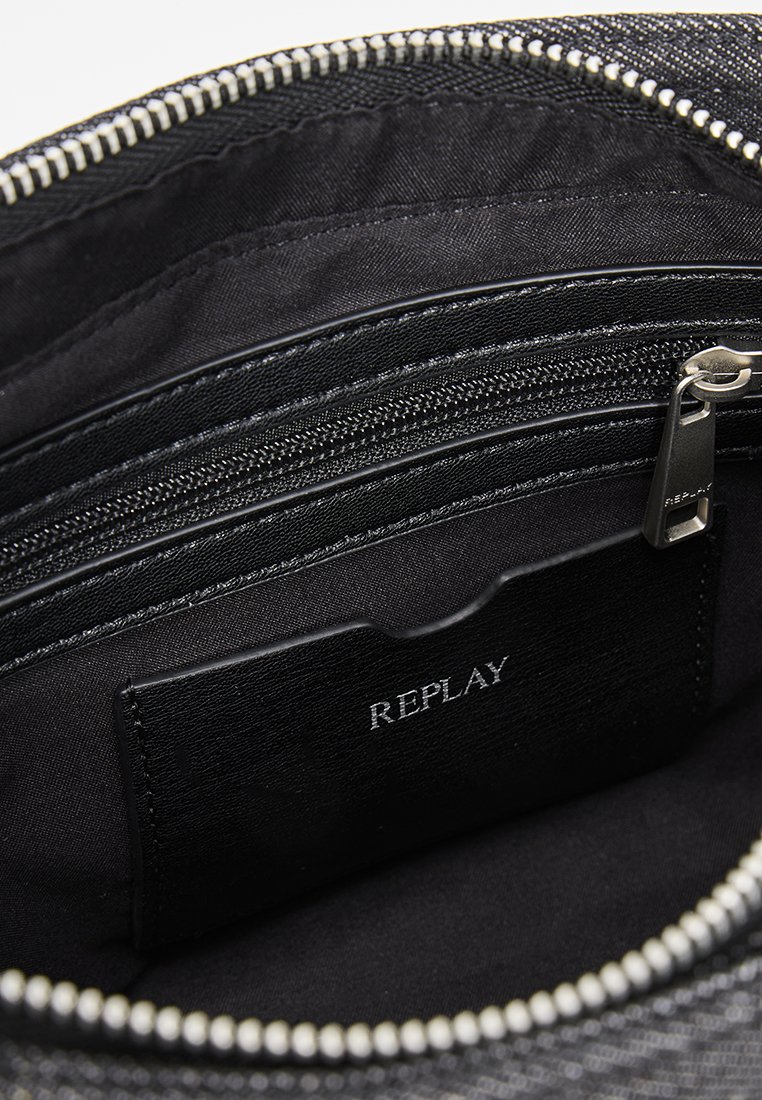 Interno di una borsa in tessuto nero con texture, dotato di molteplici scomparti con zip e un'etichetta che riporta la scritta "REPLAY" su una superficie nera liscia.