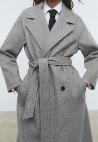 Grijze wollen blendjas met een strikceintuur, notched revers, knoopsluiting en zijzakken. Heeft een zachte textuur en een losse pasvorm.