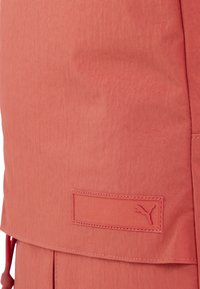 Tessuto di un rosa corallo con una texture liscia, caratterizzato da un'etichetta con logo ricamata e un design con tasca laterale.