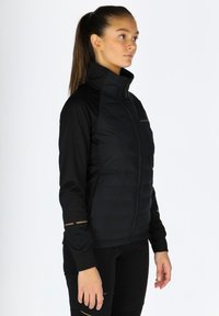 Giacca sportiva nera con parte anteriore imbottita, maniche lisce, colletto alto e dettagli dorati sui polsini. Chiusura con zip, design aderente.
