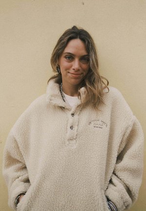 Młoda kobieta o jasnobrązowych włosach, uśmiechająca się, ubrana w beżowy polar, stojąca na tle jednolitej jasnokolorowej ściany.