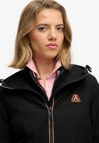 Chaqueta negra con cremallera, capucha y logo con acentos en naranja. Superpuesta sobre una camisa de color rosa claro con cuello de botones. Textura suave, diseño moderno.