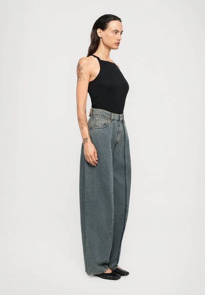 Femeie cu păr închis la culoare, îmbrăcată cu un top negru fără mâneci, jeans gri cu talie înaltă și croială largă, pantofi negri, stând de profil pe un fundal alb simplu.