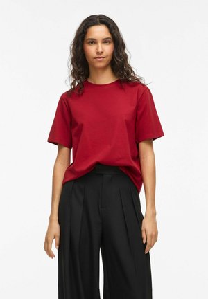 Femme aux cheveux bouclés portant un t-shirt rouge uni à manches courtes rentré dans un pantalon noir plissé taille haute, debout devant un fond blanc.
