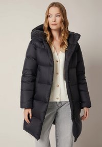 Czarna puchowa kurtka z kapturem, pikowany design i zamek błyskawiczny z przodu. W zestawie z kremowym kardiganem i jasnoszarymi dżinsami. Gładka tekstura.